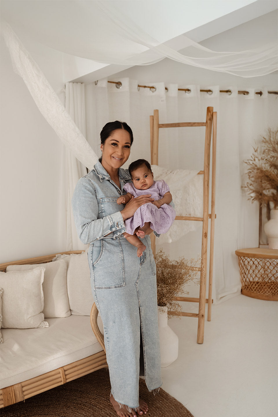 Woman holding a baby in a room with white walls, a wooden ladder, and a couch.